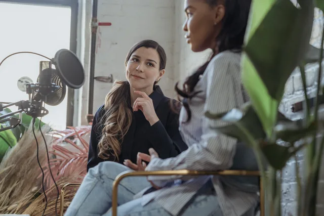 Two women engaging in a podcast conversation indoors with microphone setup. Downloads, listeners and ad impressions explained