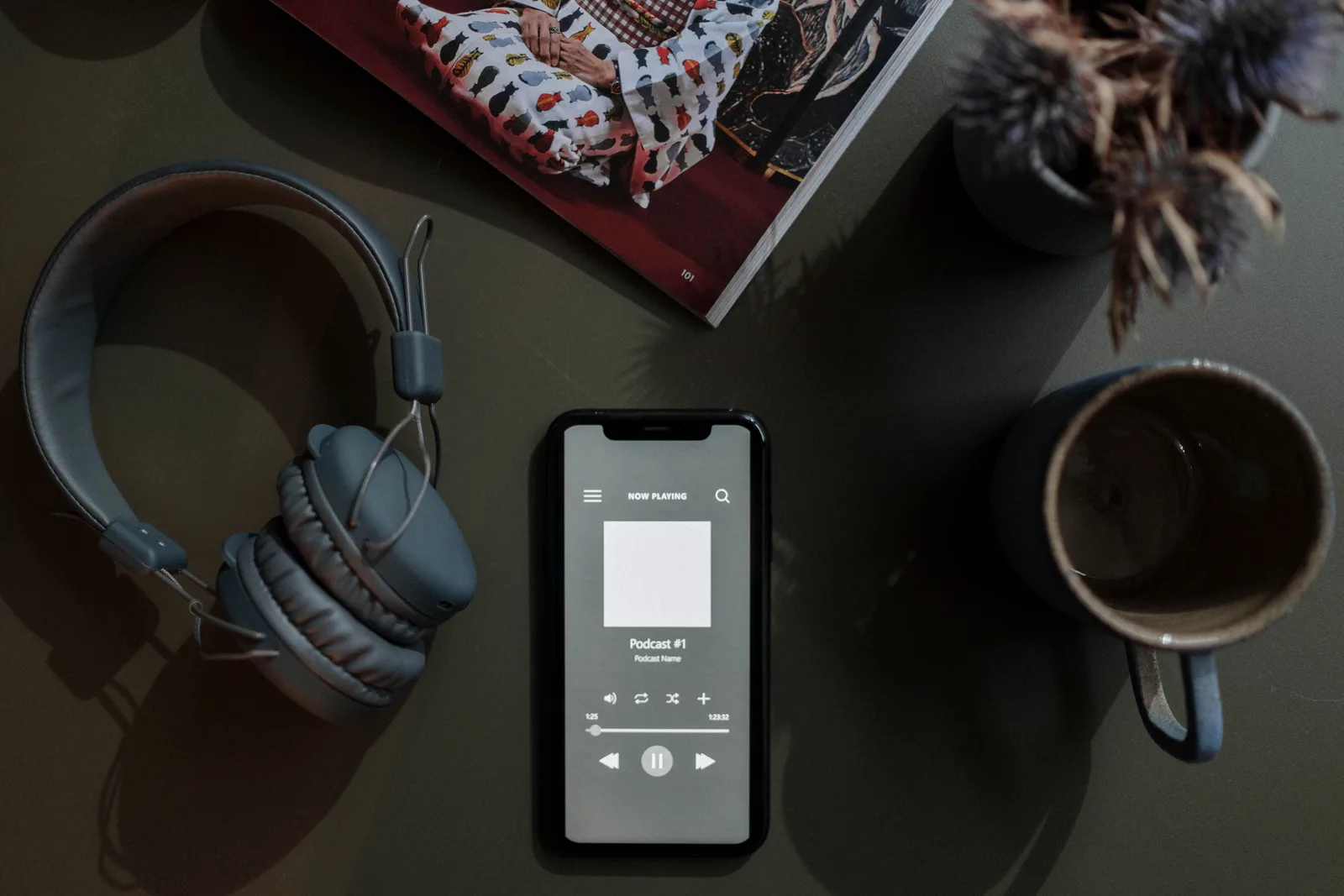 A flat lay image of a smartphone playing a podcast next to headphones and a mug.